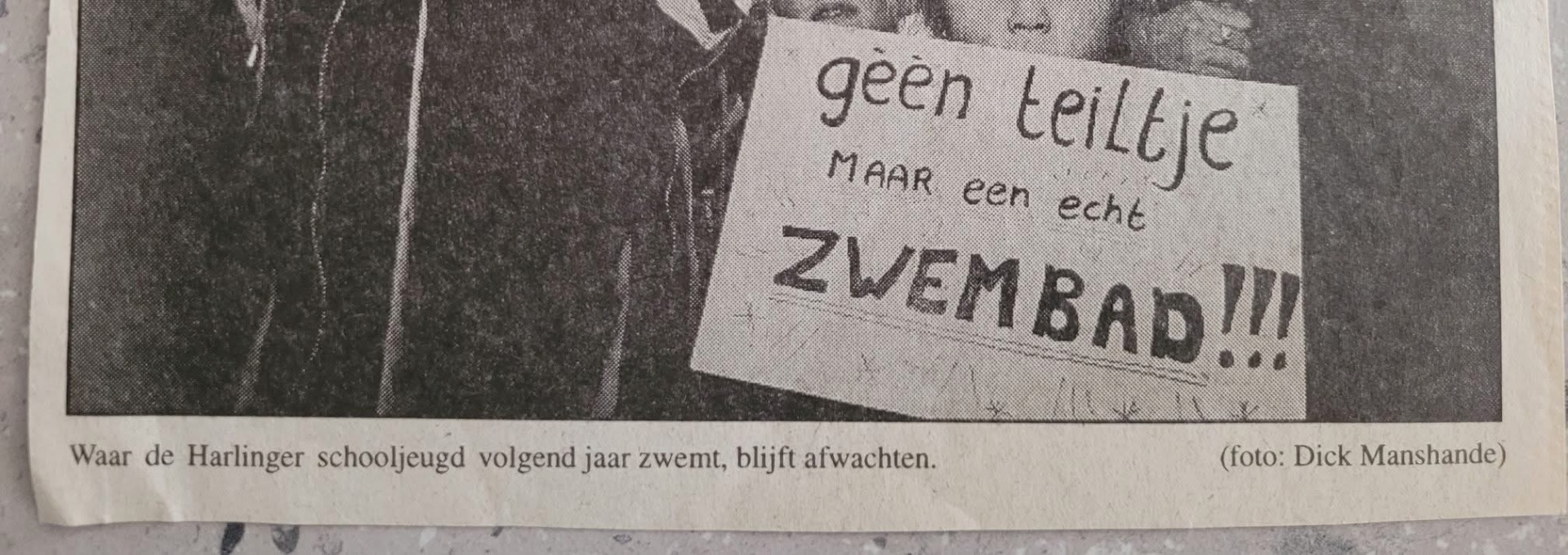 Historisch krantenknipsel met een bord waarop staat: geen teiltje maar een echt zwembad. Onder de foto de tekst: Waar de Harlinger schooljeugd volgend jaar zwemt, blijft afwachten.
